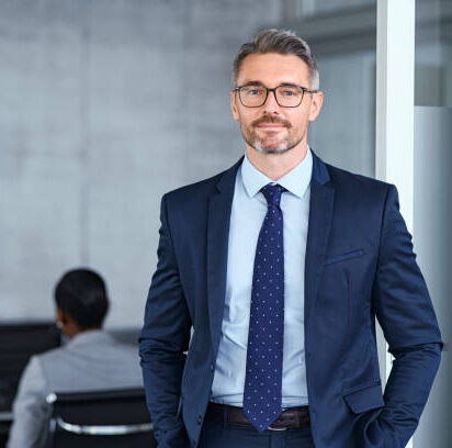 Portrait of handsome mid adult business man standing in modern office. Successful mature entrepreneur in formal clothing looking at camera with satisfaction. Confident man in suit with eyeglasses and beard standing with hands in pocket and looking at camera in office.