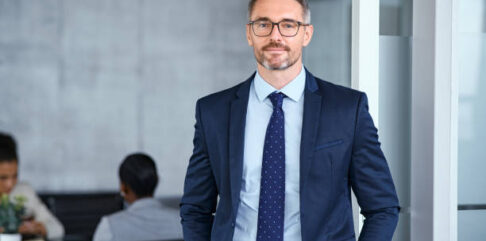 Portrait of handsome mid adult business man standing in modern office. Successful mature entrepreneur in formal clothing looking at camera with satisfaction. Confident man in suit with eyeglasses and beard standing with hands in pocket and looking at camera in office.