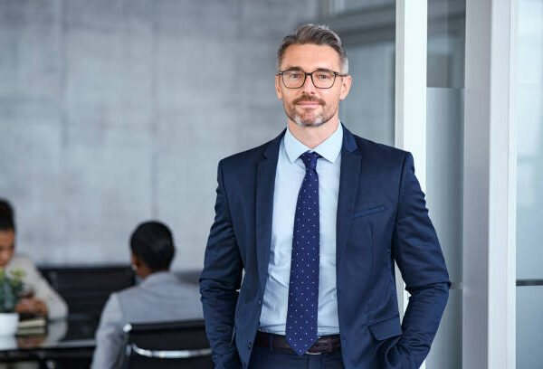 Portrait of handsome mid adult business man standing in modern office. Successful mature entrepreneur in formal clothing looking at camera with satisfaction. Confident man in suit with eyeglasses and beard standing with hands in pocket and looking at camera in office.